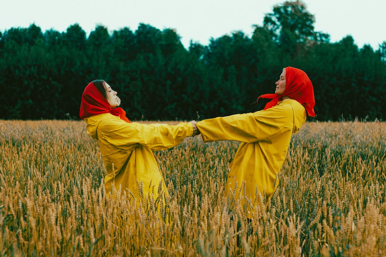 Women wearing yellow raincoats and red headscarves dance joyfully in a wheat field.