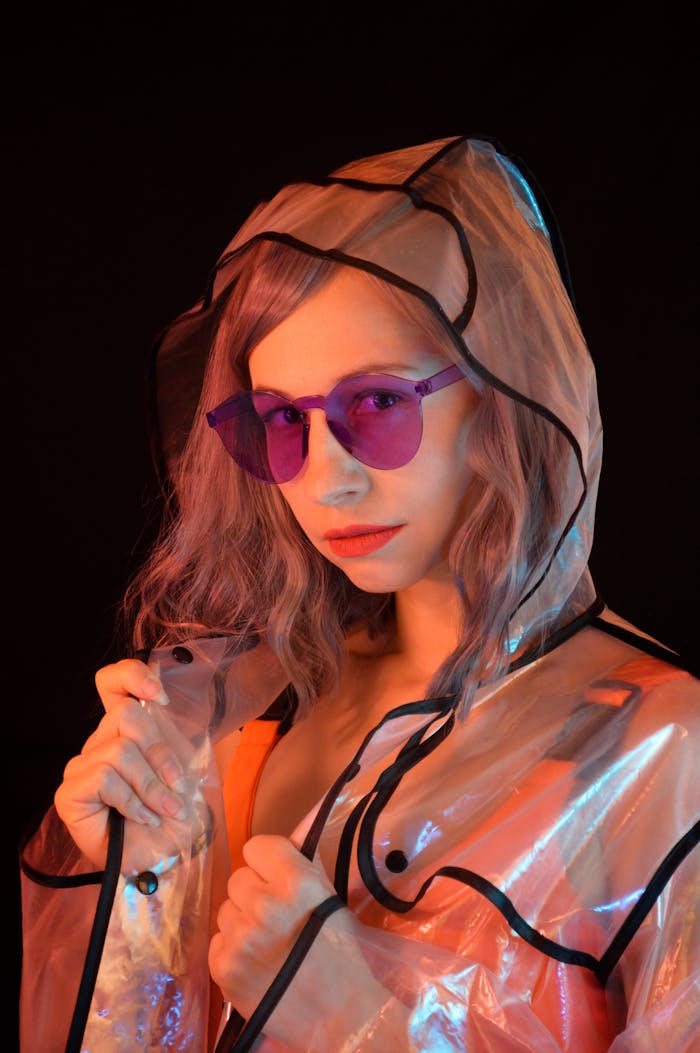 Stylish female model in trendy purple sunglasses and transparent raincoat standing in studio and looking at camera against black background
